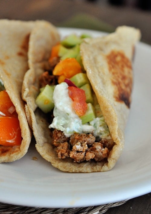 A flatbread taco full of meat, diced cucumbers, and Greek sauce on a white plate.