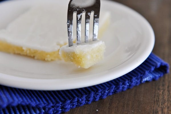 A piece of white Texas sheet cake on a white plate with a fork taking a bite out.