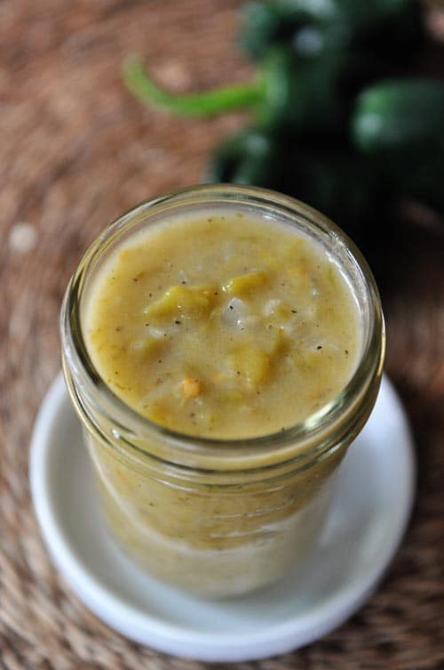 A top-view of a mason jar full of green enchilada sauce, with jalapeños in the background.