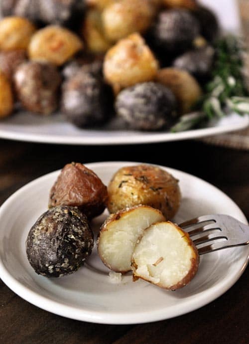 White plate with four small cooked potatoes with one cut in half and a white platter with more potatoes behind it.