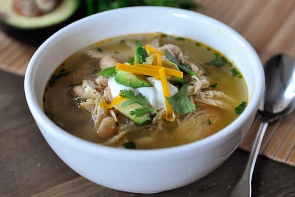 White bowl of white bean chicken chili with sour cream, diced avocado, and cheese.