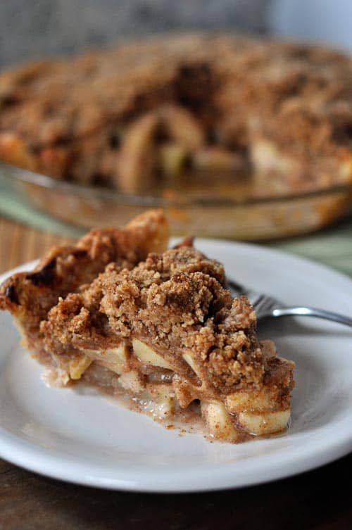 A slice of apple crumb pie on a white plate with the rest of the pie in the background.