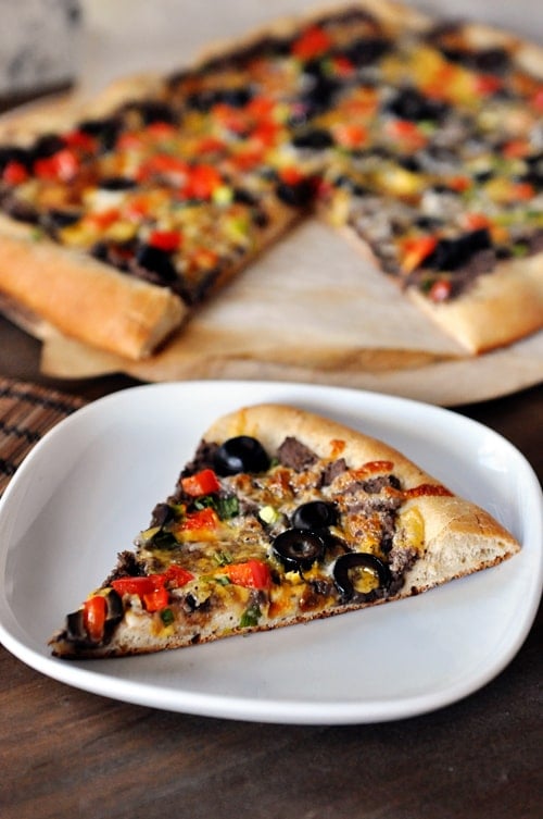 A slice of black bean veggie pizza on a white plate with the rest of the pizza behind it.