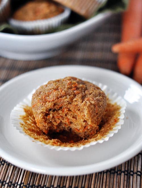 A carrot cake muffin with one bite taken out on a white liner that's been peeled off the muffin. 