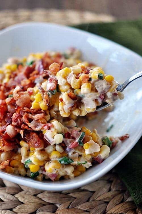 A bowl of creamy confetti corn and bacon with a spoon taking one big bite out. 