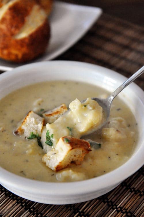 white bowl of roasted cauliflower soup with croutons on top and a spoon taking a bite out