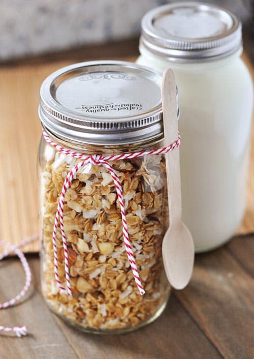 A mason jar filled with coconut granola with a spoon and ribbon tied around it, and a mason jar filled with yogurt behind it.