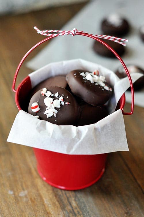 Homemade chocolate-dipped peppermint patties, with crushed candy canes on top of each patty, all in a small red bucket. 