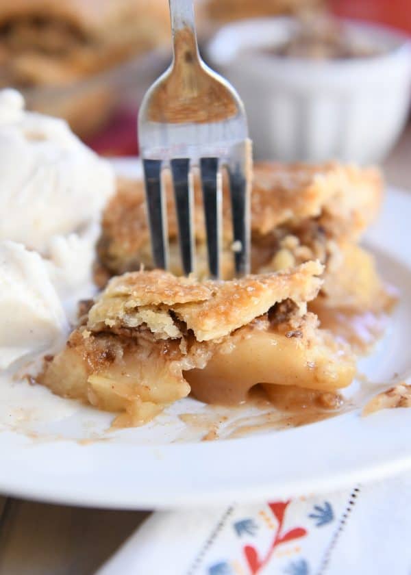 Fork cutting off bite of toffee caramel apple pie on white plate with vanilla ice cream.
