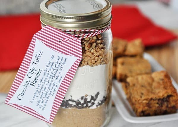 a glass mason jar with dry ingredients for chocolate chip toffee blondies and cooked blondies on a platter on the side