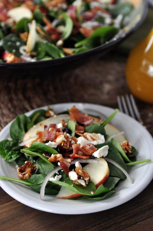 A plate of spinach salad with chopped nuts, onions slices, bacon, apple slices, and feta on top. 