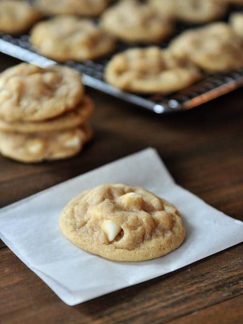 A white chocolate madadamia nut cookie on a piece of parchment paper, with other macadamia cookies on a cooling rack and stacked up behind in the background.