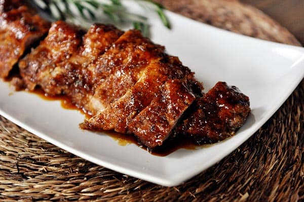 A sliced, glazed pork tenderloin, with a sprig of rosemary, all on a white platter.