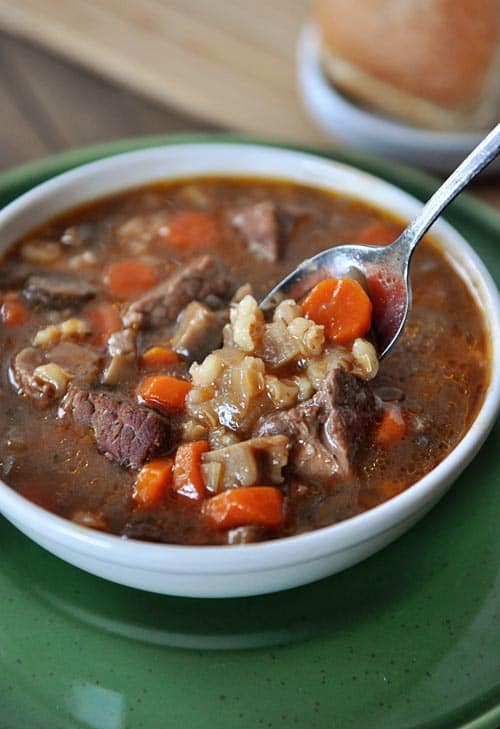 A white bowl full of beef, barley, and carrot stew with a spoon taking a bite out.