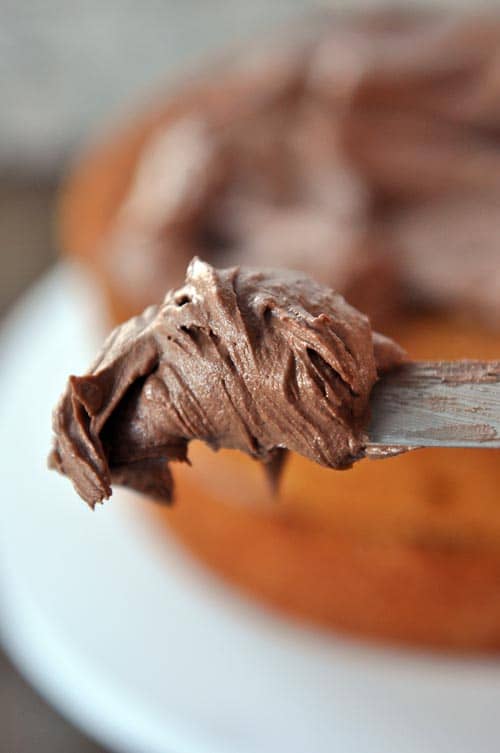 A metal spatula with chocolate frosting and a cake that is getting frosted in the background.
