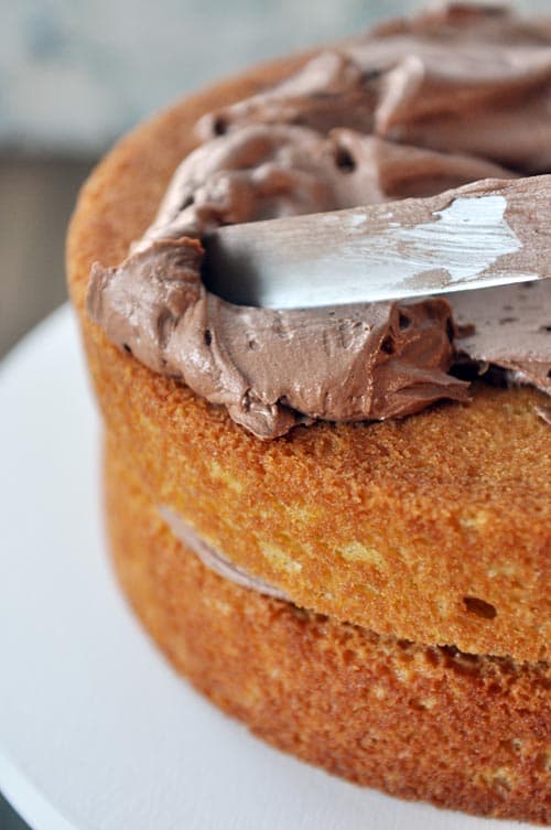 A double decker cake getting frosted with chocolate frosting.