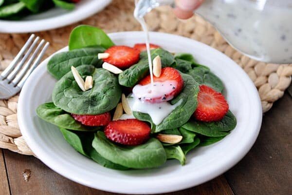 Someone pouring poppy seed dressing over a spinach strawberry salad.