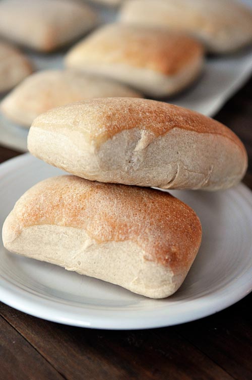 Two golden brown rectangular shaped italian rolls stacked on top of each other on a white plate, with the rest of the rolls on a cookie sheet in the background.