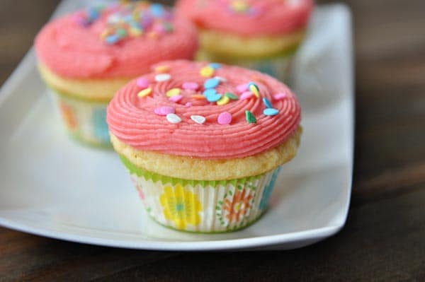 Yellow cupcakes with pink frosting and sprinkles on a white platter.