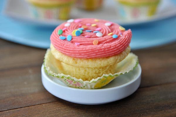 a yellow cupcake with pink frosting and sprinkles sitting in a white ramekin
