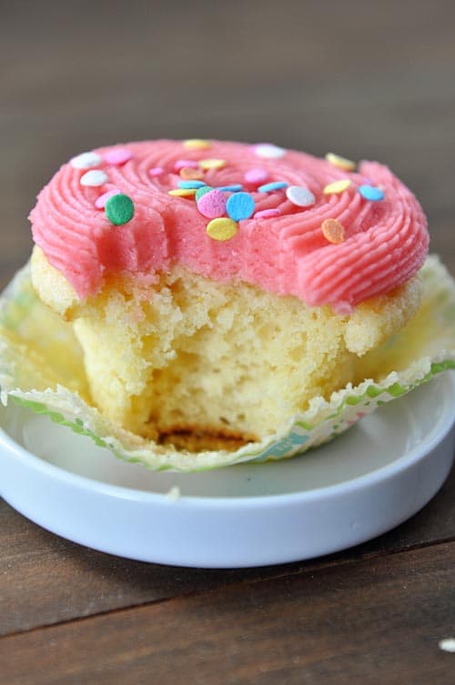 A yellow cupcake with pink frosting and sprinkles on a small white dish. The cupcake has a bite taken out. 