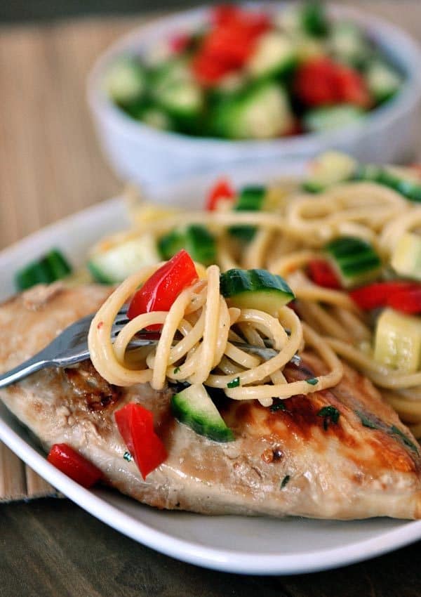 A white plate with grilled chicken topped with a spaghetti cucumber tomato mixture.