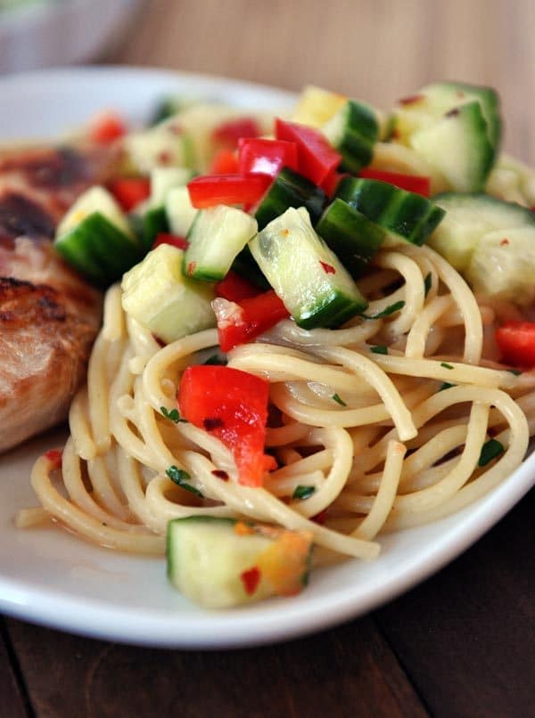 a white plate with cooked spaghetti noodles topped with chopped cucumbers and tomatoes