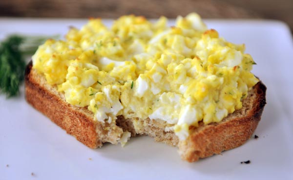 Open faced egg salad sandwich with a bite taken out on a white plate.