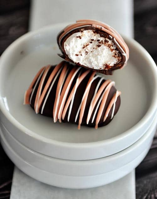 Two chocolate covered and chocolate drizzled marshmallow eggs on white plates. The top marshmallow egg has a bite taken out. 