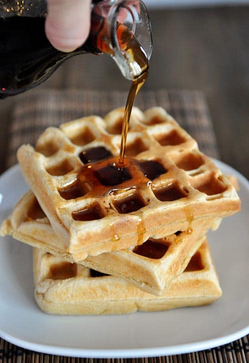 A hand pouring syrup over a stack of thick waffles.