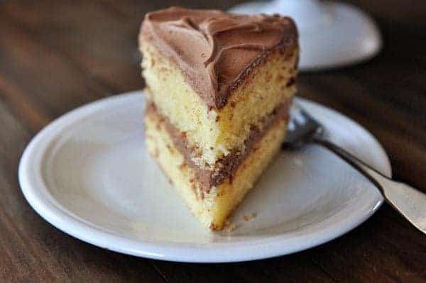A piece of yellow cake with chocolate frosting on a white plate, with a fork next to the piece of cake.