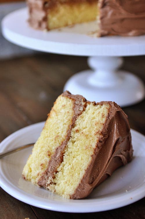 A piece of yellow cake with chocolate frosting on a white plate, with the rest of the cake in the background on a cake stand.