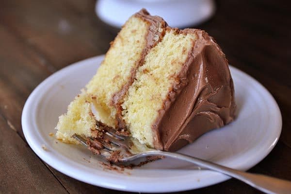 A fork getting a bite out of a piece of yellow cake with chocolate frosting on a white plate. 