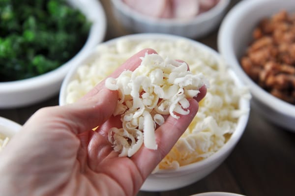 White bowls full of pizza toppings and someone holding a handful of cheese over the cheese bowl.