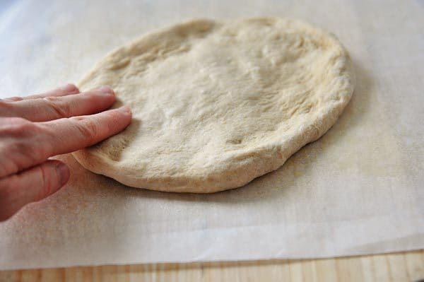 Uncooked pizza dough on a piece of parchment paper, being flattened to a crust. 