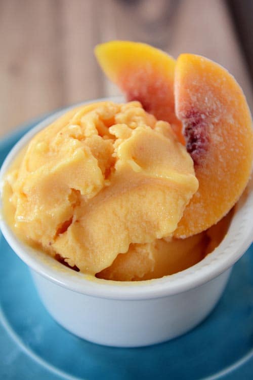 A small white ramekin with scoops of fresh peach frozen yogurt and a few frozen peach slices. 