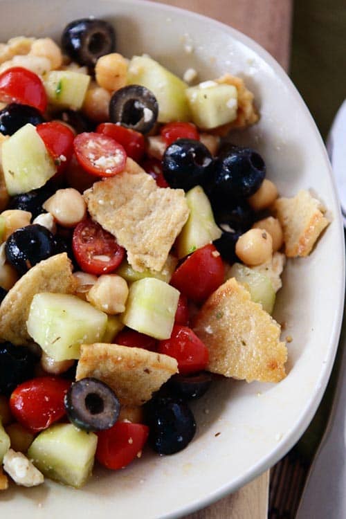 Top view of a white bowl of greek pita salad filled with pitas, cucumbers, olives, tomatoes, and chickpeas. 