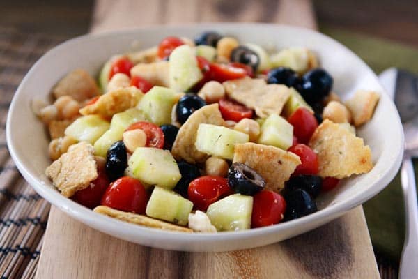 A white bowl full of pita cubes, olives, cucumbers, olives, and tomatoes with a light dressing.