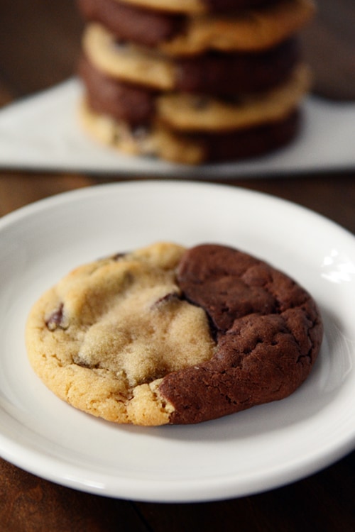 A cookie with half brownie, half chocolate chip cookie on a white plate, and a stack of the same cookies in the background.
