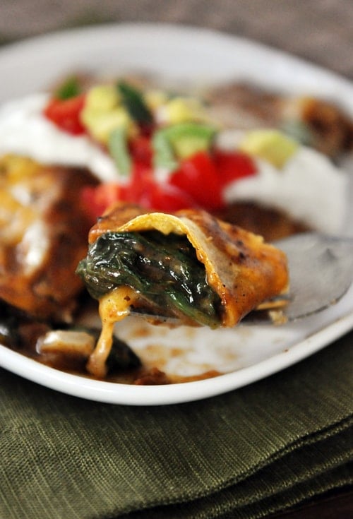 A bite of a spinach enchilada on a fork with the rest of the enchilada and toppings on a white plate below it.