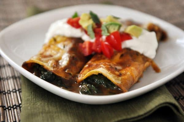 Two spinach enchiladas topped with sour cream, tomatoes, and avocados on a white plate. 