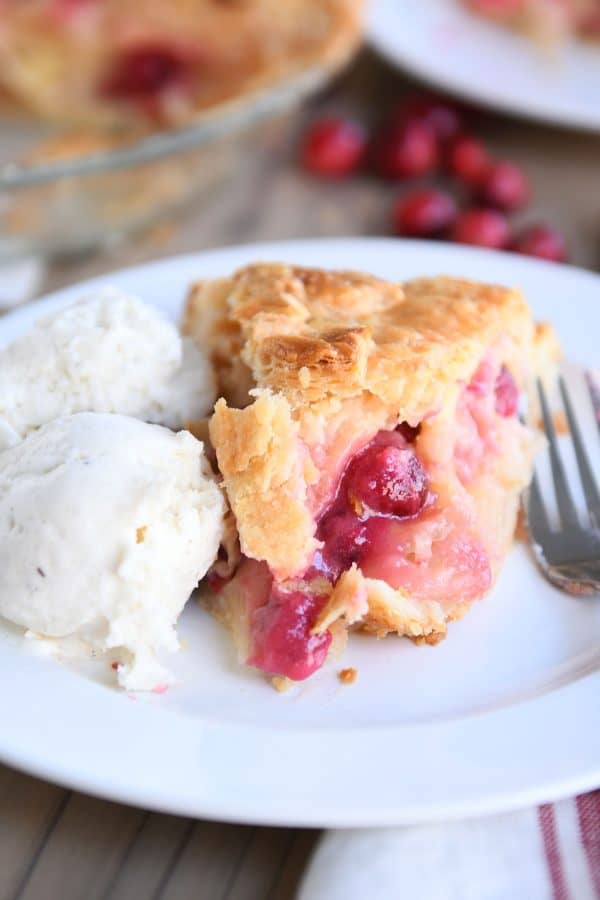 Slice of apple cranberry pie on white plate with ice cream.