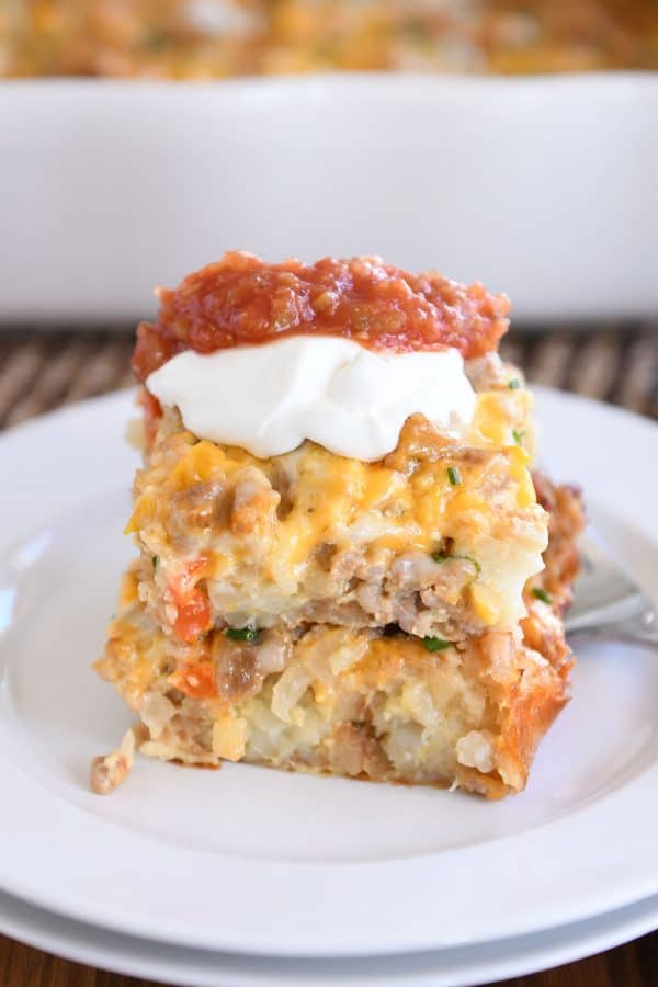 Two squares of tater tot breakfast casserole stacked on each other on white plate with sour cream and salsa.