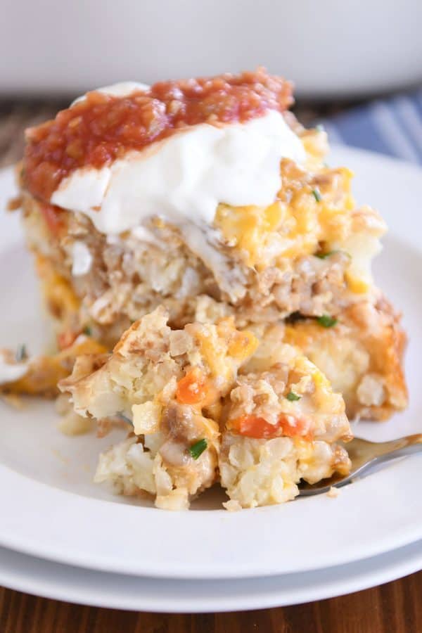 Two squares of tater tot breakfast casserole stacked on each other on white plate with sour cream and salsa and fork taking a bite