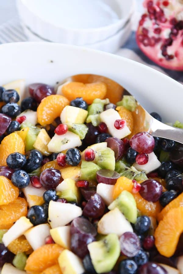 White bowl and spoon with grapes, blueberries, pears, kiwi, oranges and pomegranate seeds for easy winter fresh fruit salad.