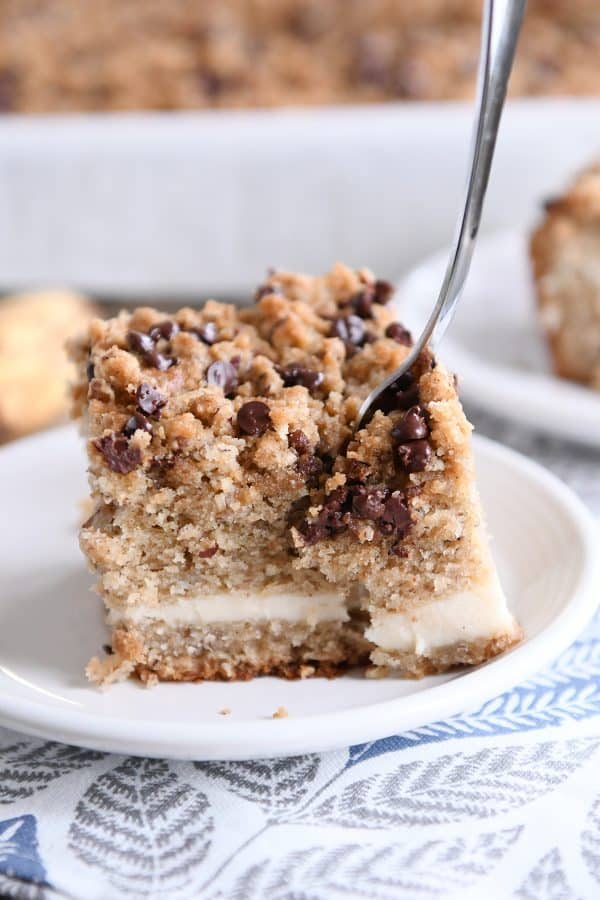 A fork taking a bite of a piece of banana cream cheese coffee cake on a white plate.