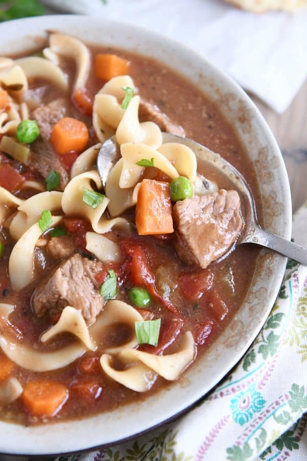 Top down view of slow cooker beef vegetable noodle soup with spoon in gray ceramic bowl.