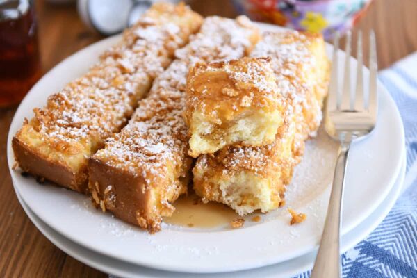 Crunchy baked french toast sticks on white plate with powdered sugar and syrup.
