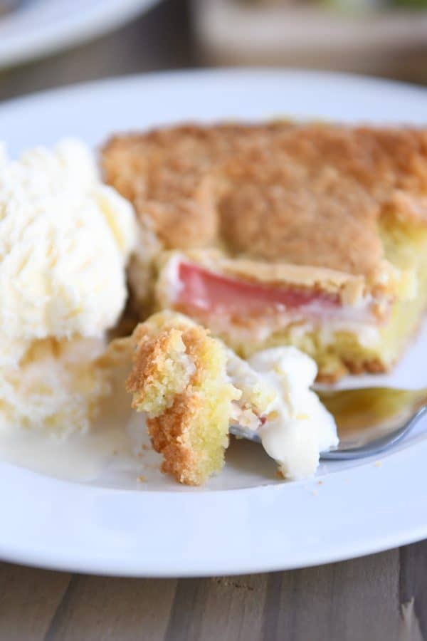 piece of rhubarb custard cake on white plate with ice cream and fork taking out a bite