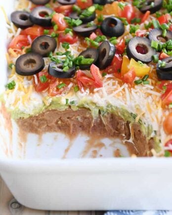 scoop taken out of 7-layer dip in white casserole dish